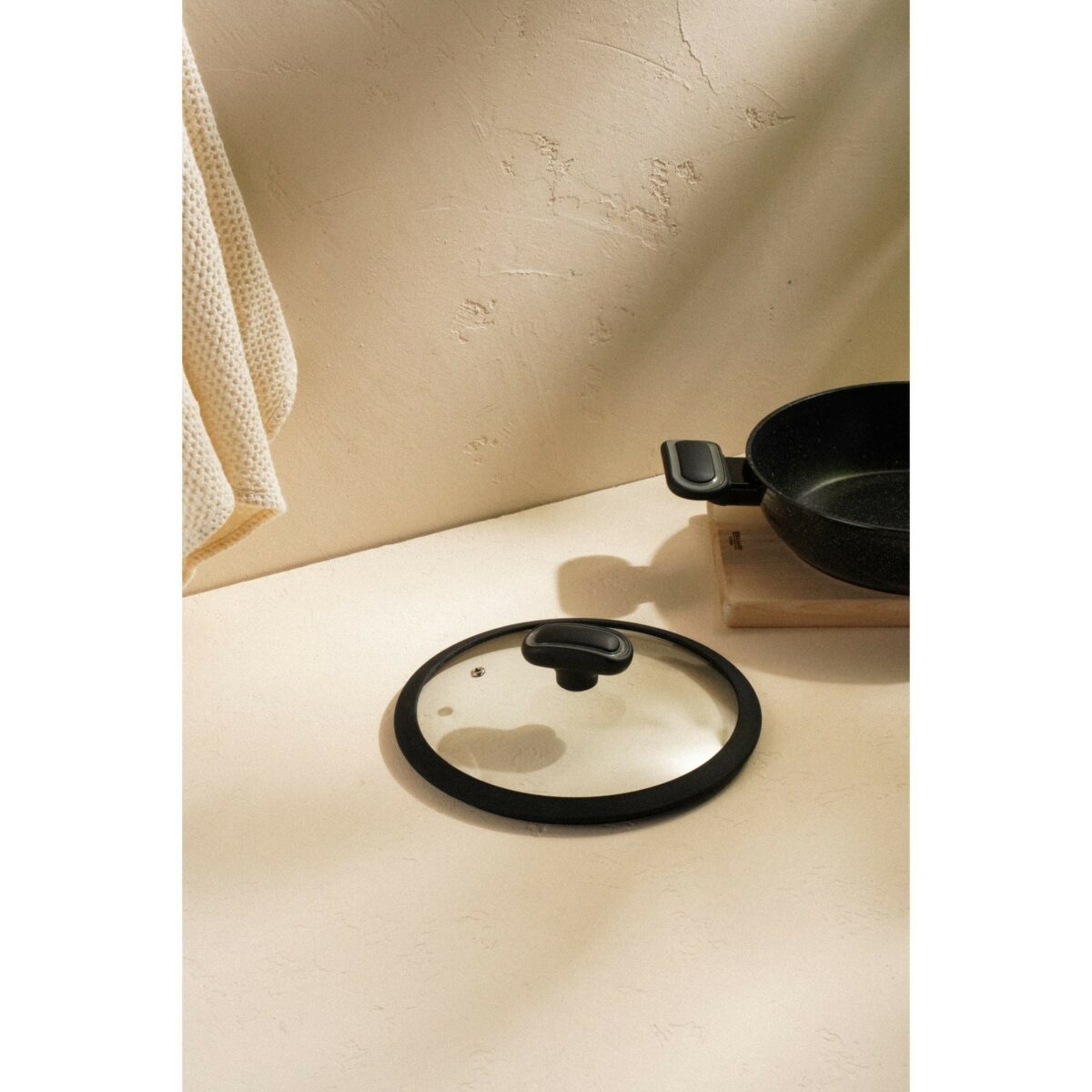 Стеклянная крышка для сковороды 22 см с силиконовым ободком и отверстием для выхода пара Универсальная стеклянная крышка с силиконовым ободком для сковороды диаметром 22 см, лежащая на светлой поверхности рядом с сковородой на деревянной подставке