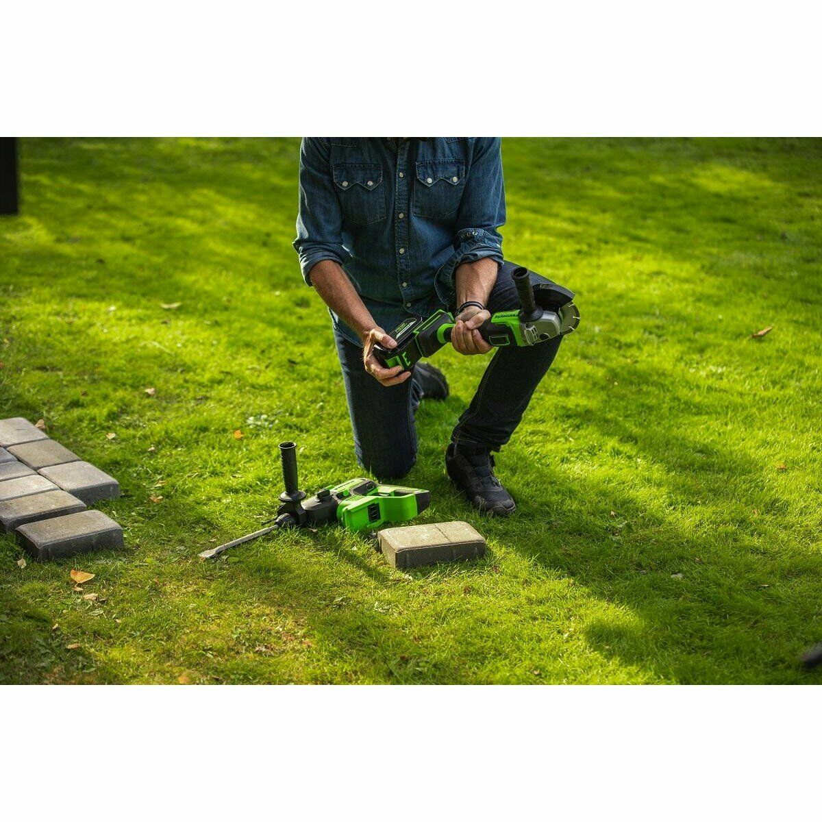 Человек на газоне работает с электрическим инструментом зеленого цвета рядом с брусчаткой