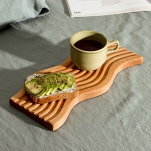 Деревянная доска для подачи с волнообразной поверхностью, хлеб с авокадо, зелёная чашка напитка и текстурный фон