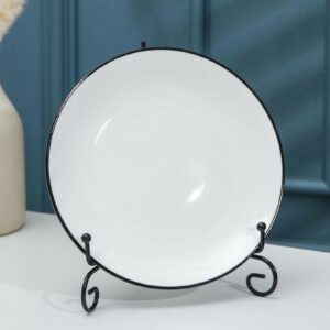 Черная прочная подставка под тарелки из металла с декоративными изгибами на белом столе, в интерьере с голубой стеной