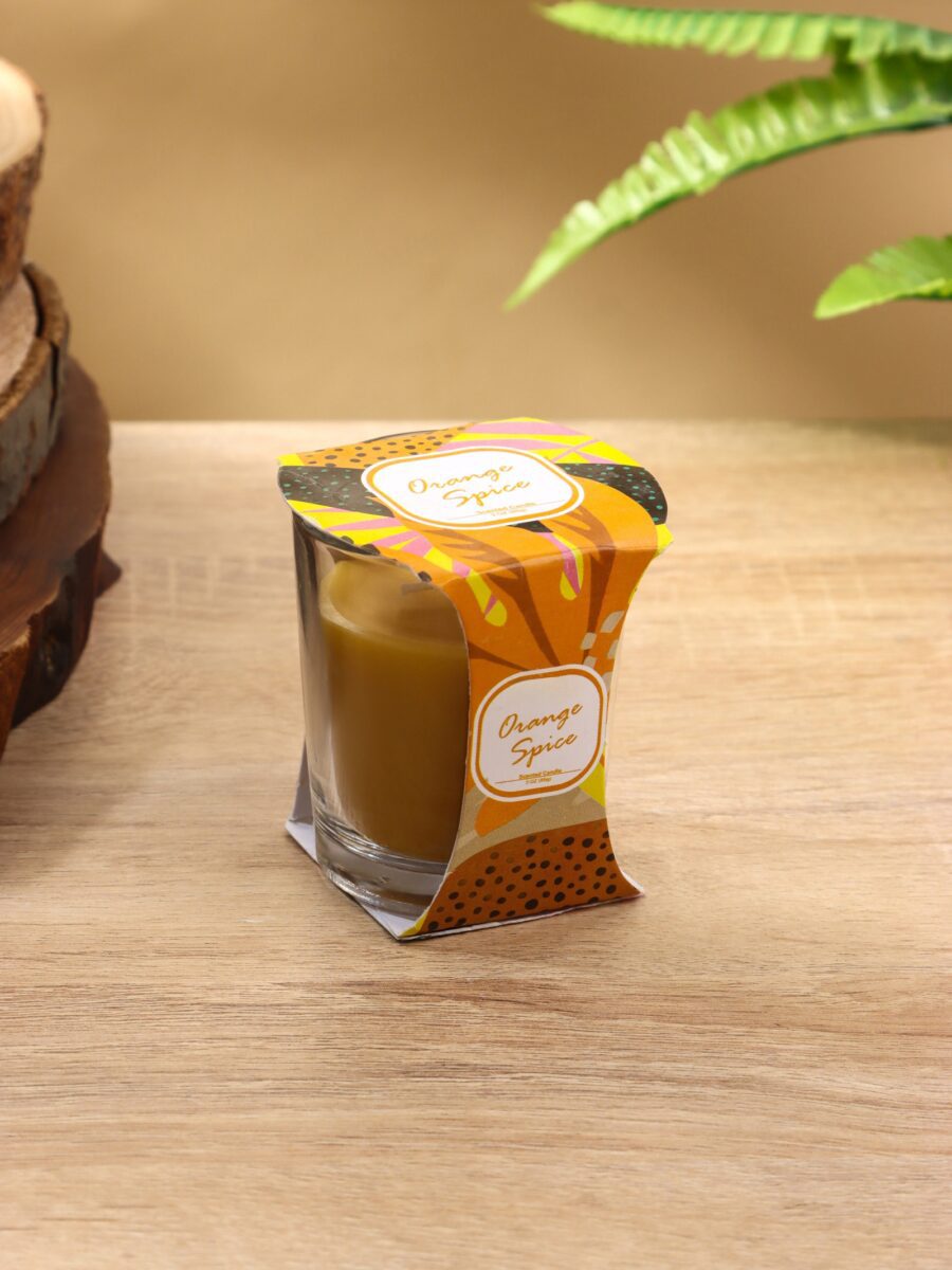 Ароматическая свеча в стеклянном стакане с насыщенным оранжевым воском и упаковкой из картона с элементами оранжевого, желтого и зеленого цветов. Подходит для украшения дома и наполнения пространства ярким ароматом апельсина и специй.